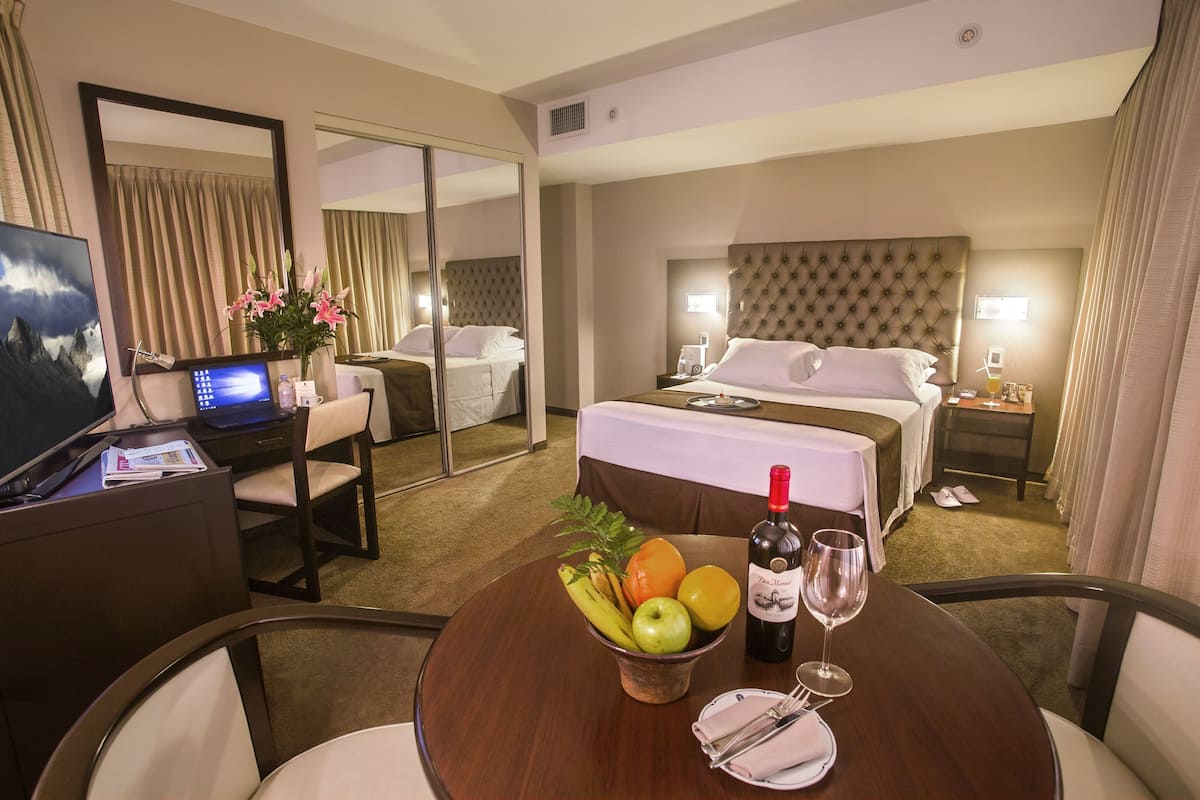 A hotel room with a bed, bedside table, a desk with a computer, and a dining table with fruit and a wine glass.