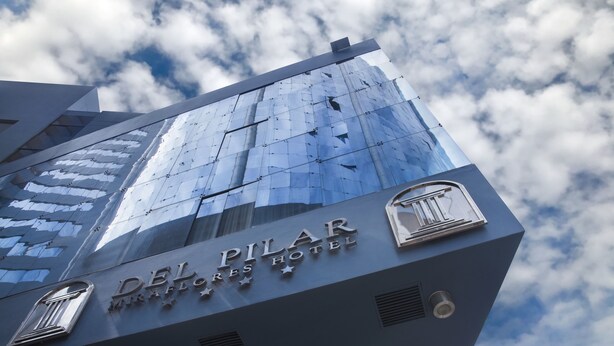 Exterior del Hotel Del Pilar con fachada de vidrio moderna que refleja el cielo y las nubes.