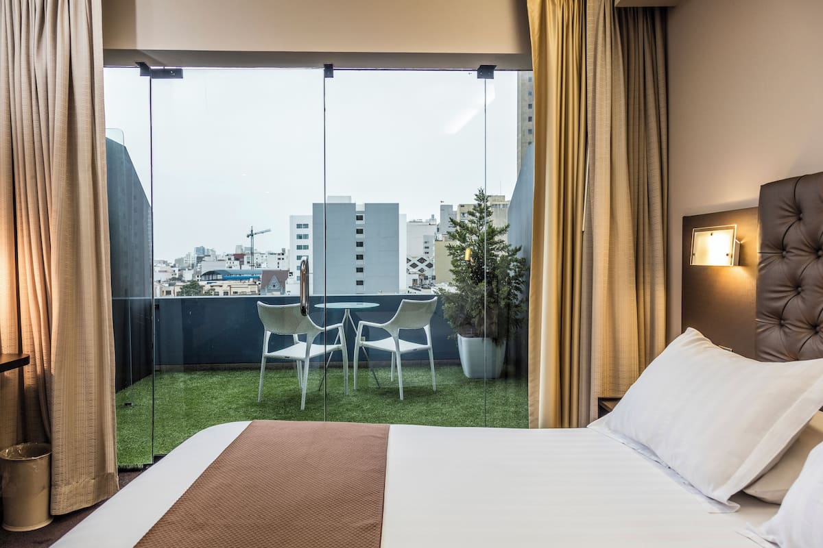A hotel room with a bed, a small balcony with a table and chairs, a potted plant, and a city view.