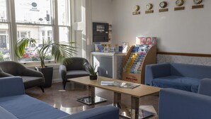 A reception area with a reception desk, blue seating, a potted plant, a window with a view of buildings, and a wall with clocks and a 'Take One' magazine stand. - Best Western Corona (London)