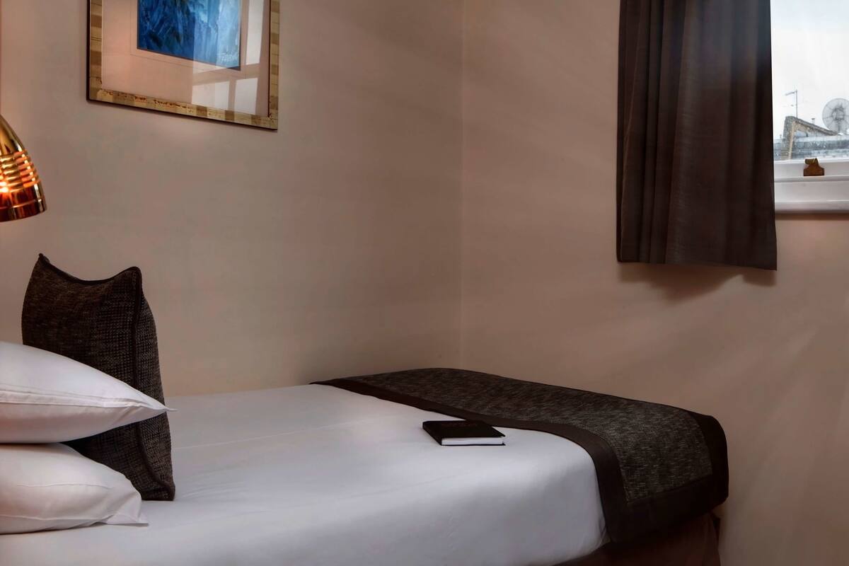 A hotel room with a bed, pillows, a bedside lamp, a window with curtains, and a framed picture on the wall.