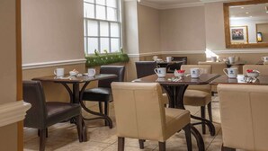 A dining area with tables and chairs, a window with blinds, a mirror, and a small plant on the windowsill. - Best Western Corona (London)