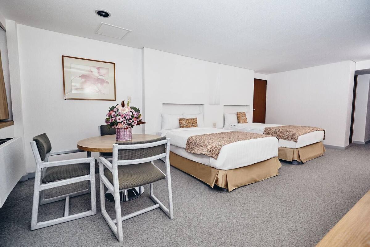 Une chambre d’hôtel avec deux lits, une table à manger avec des chaises, un vase de fleurs et un tableau encadré au mur.