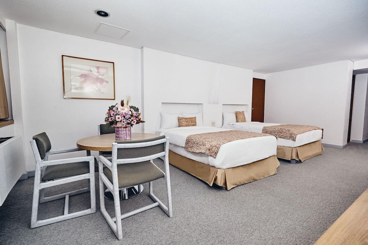 A hotel room with two beds, a dining table with chairs, a vase of flowers, and a framed picture on the wall.