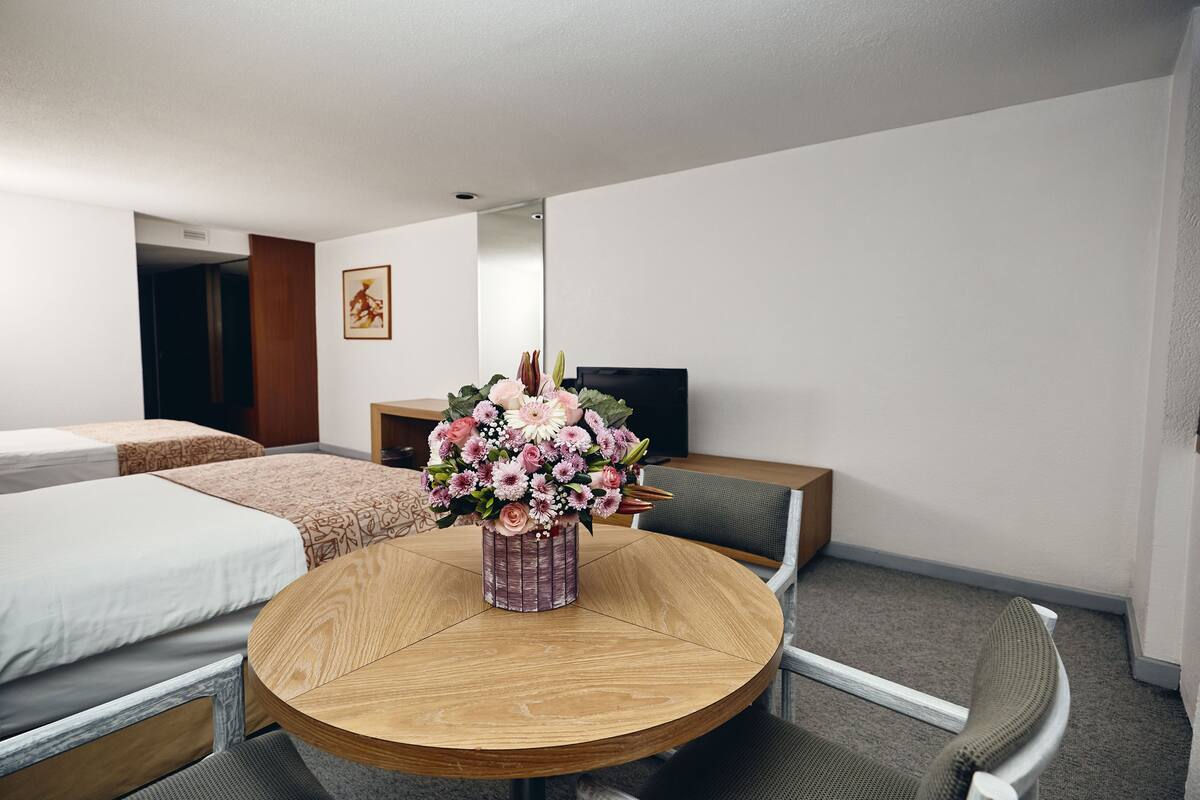 Une chambre d’hôtel avec deux lits, une table ronde en bois sur laquelle est posé un vase de fleurs, une télévision et une chaise.