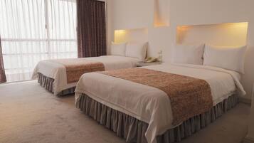 Habitación de hotel con dos camas, un ventanal grande con cortinas translúcidas, una mesita de noche con lámpara y un teléfono.