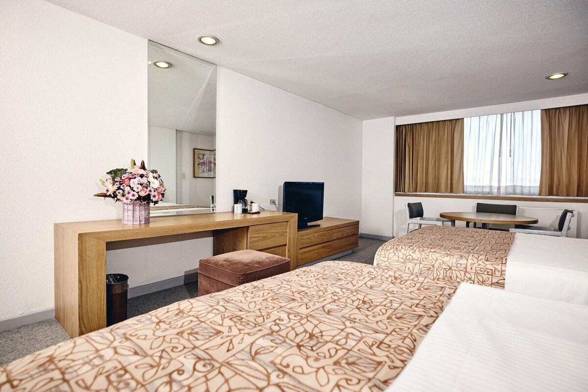 A hotel room with two beds, a desk with a TV, a mirror, and a window with curtains.