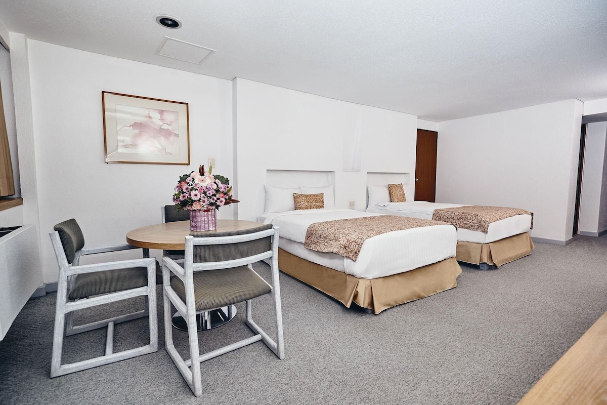 Habitación de hotel con dos camas, una mesa de comedor con sillas, un florero y un cuadro en la pared.