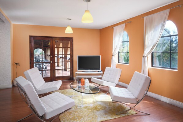 Una sala de estar con una mesa de centro de vidrio, sillas blancas acolchadas, un televisor de pantalla plana y ventanas grandes con cortinas.