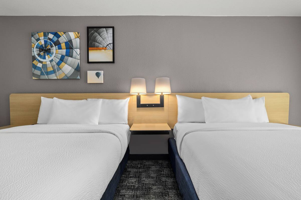 A hotel room with two beds, a headboard, two wall-mounted lamps, and two artworks on the wall.