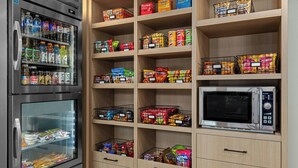 A kitchenette with a refrigerator, microwave, and shelves stocked with snacks and beverages.
