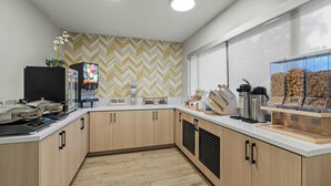 A modern kitchen with wooden cabinets, a microwave, a coffee machine, and a display case with snacks.