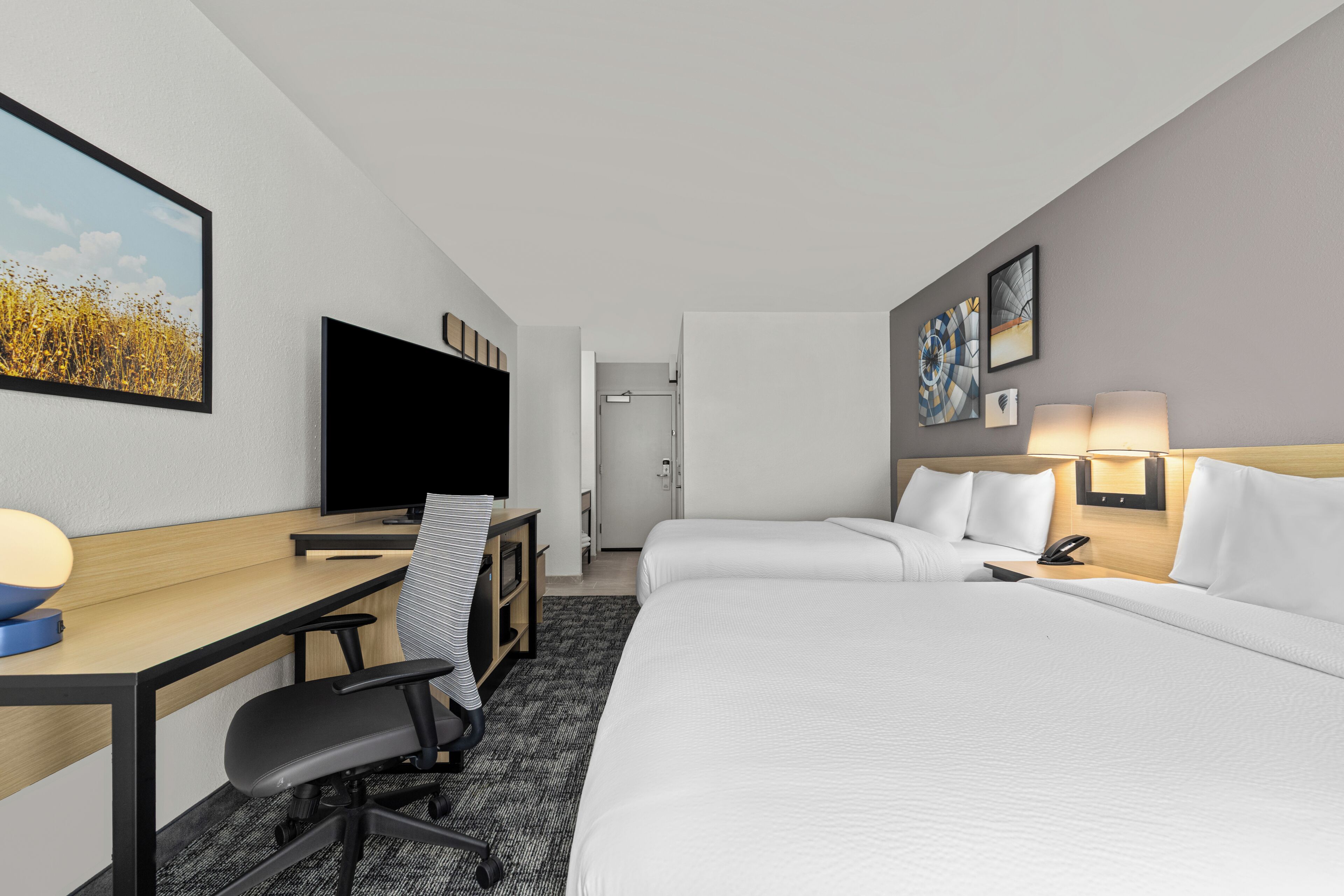 A hotel room with a desk, chair, bed, TV, and artwork on the walls.