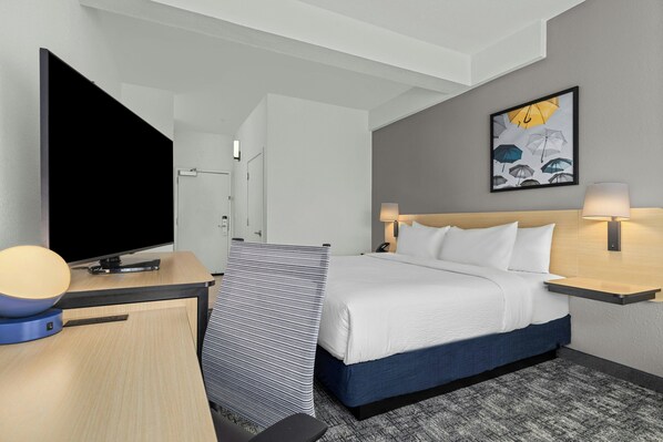 A hotel room with a bed, a desk with a flat-screen TV, a wall-mounted artwork, and a bedside table with a lamp.