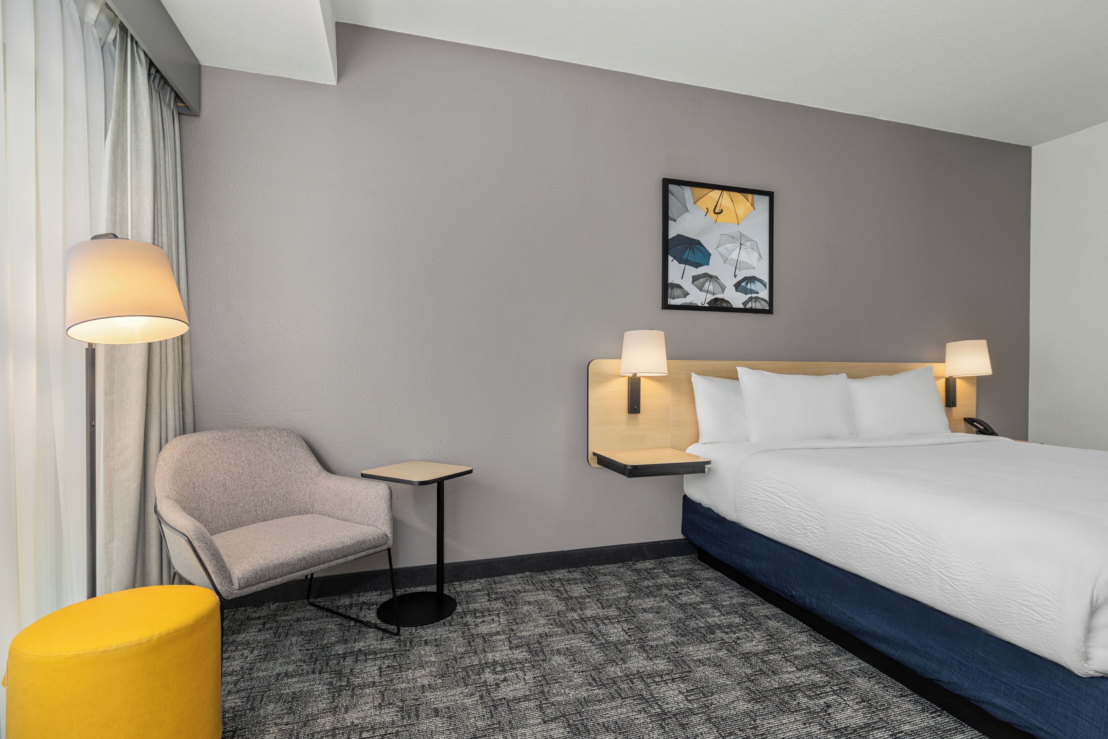 A hotel room with a bed, two bedside lamps, a chair, a small table, and a painting on the wall.