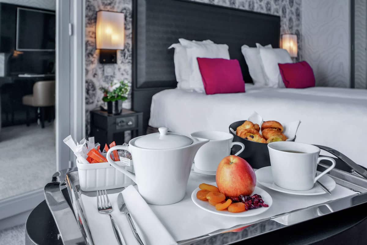 A hotel room with a neatly made breakfast tray, a bed with white and pink pillows, and a nightstand with a lamp.