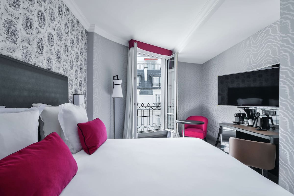 A hotel room with a bed, a desk, a chair, a TV, and a balcony with a view of buildings.