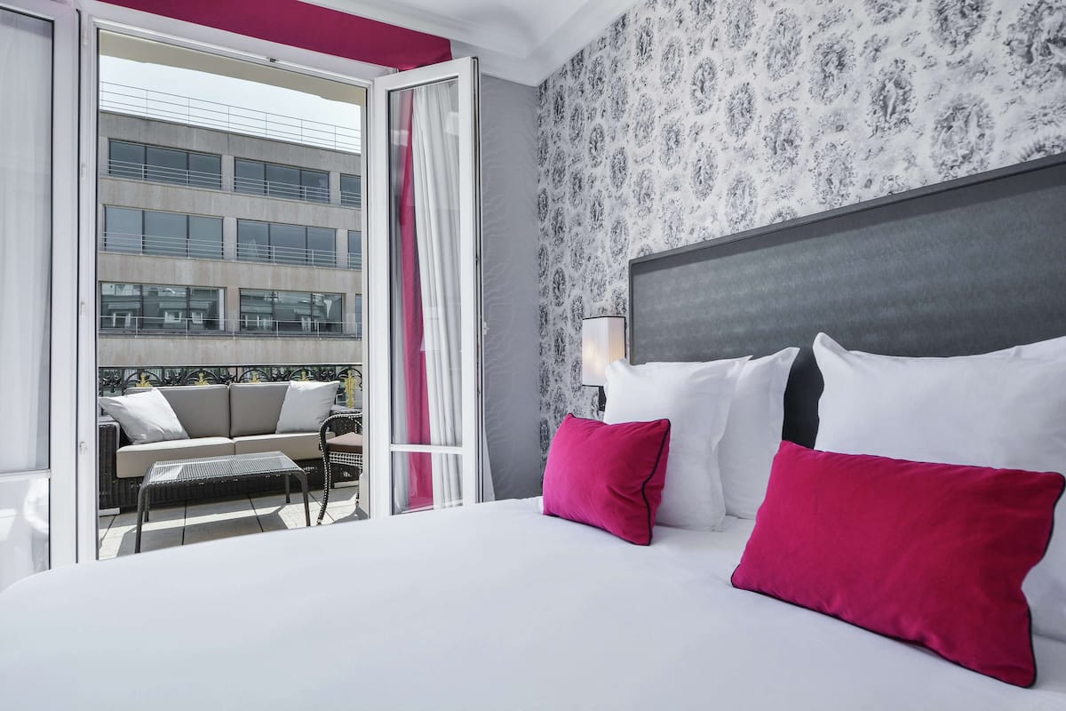 A hotel room with a bed, a balcony with seating, and a view of a building.