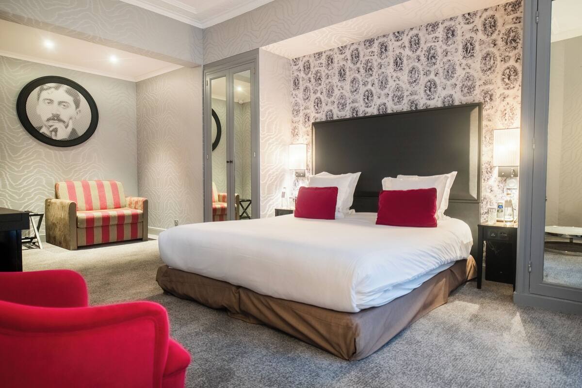 A hotel room with a large bed, a red chair, a small table, and a mirror.