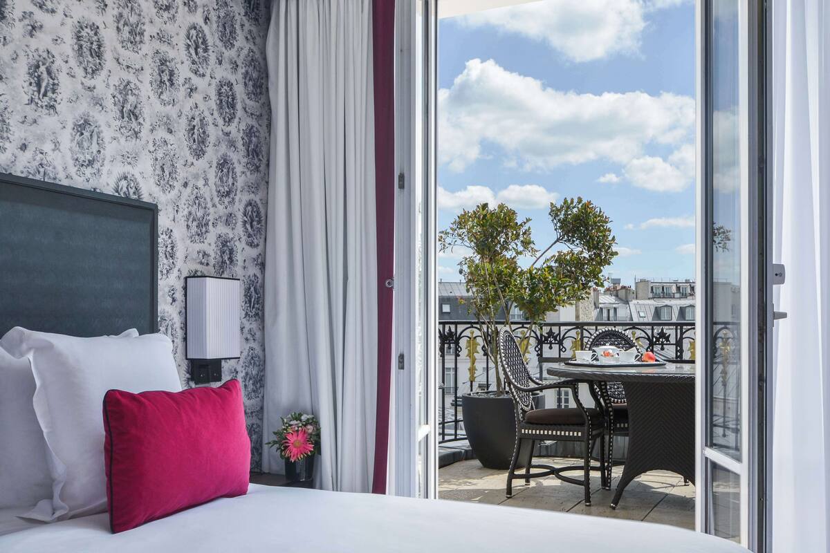 A hotel room with a bed, a red pillow, a nightstand with a lamp, a balcony with a table and chairs, and a view of the city.