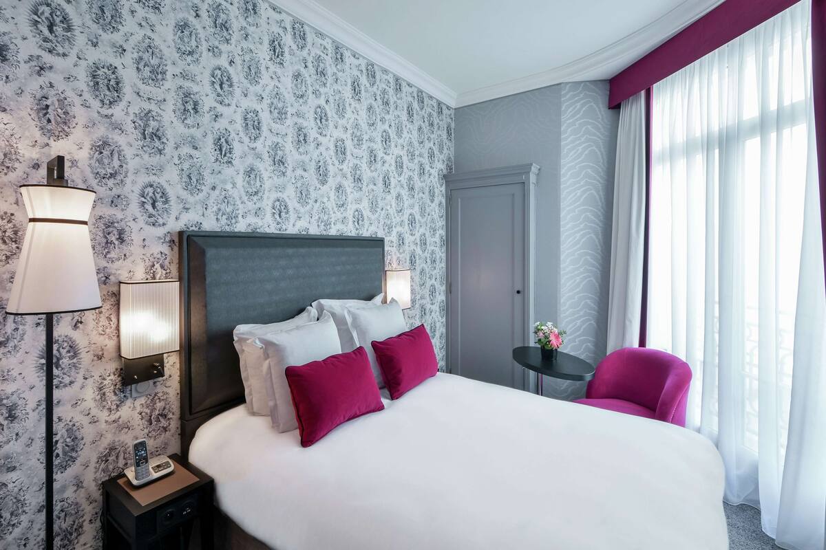 A hotel room with a large bed, a nightstand with a lamp, a small table with a flower vase, and a chair.