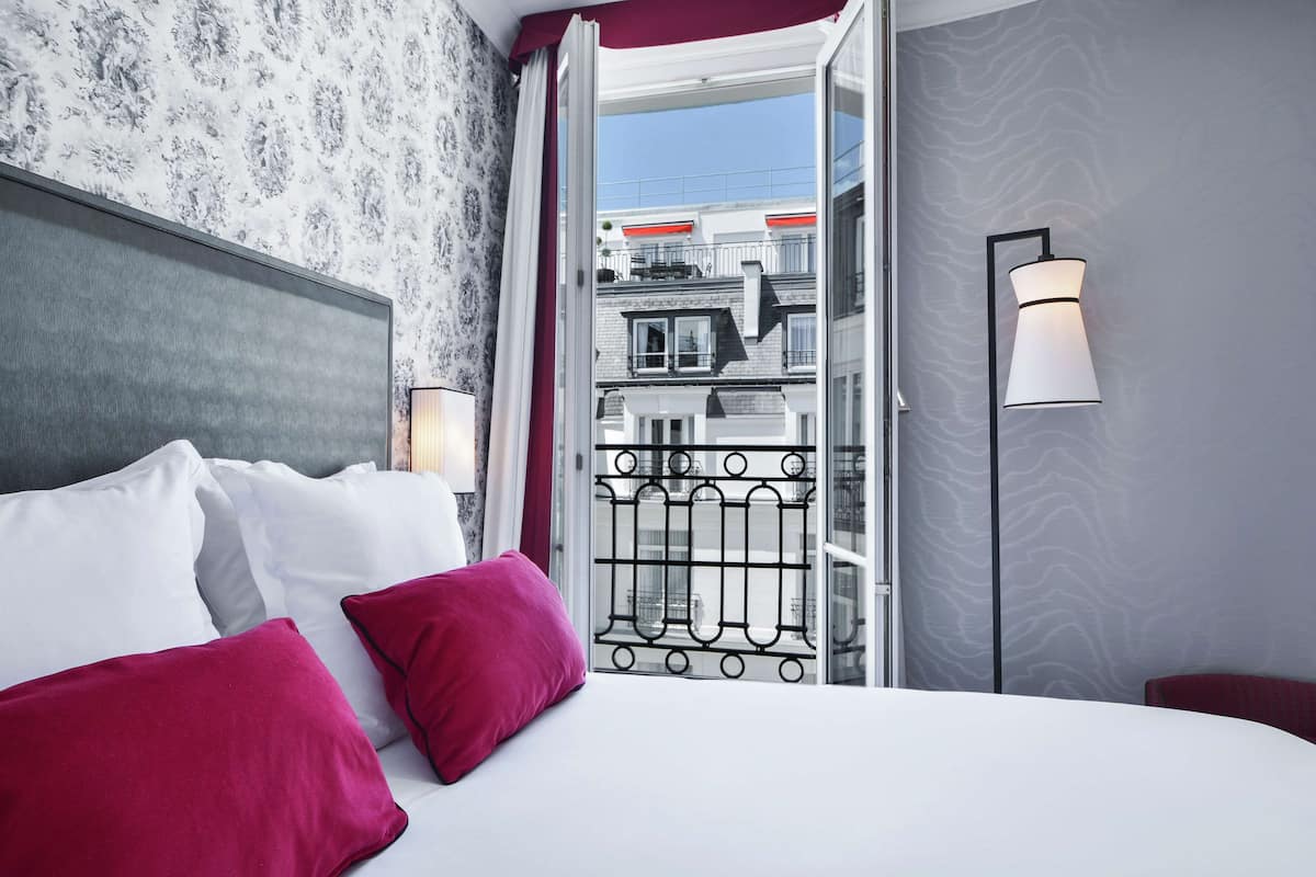 A hotel room with a bed, pillows, a nightstand, a lamp, and a view of the city through a window.