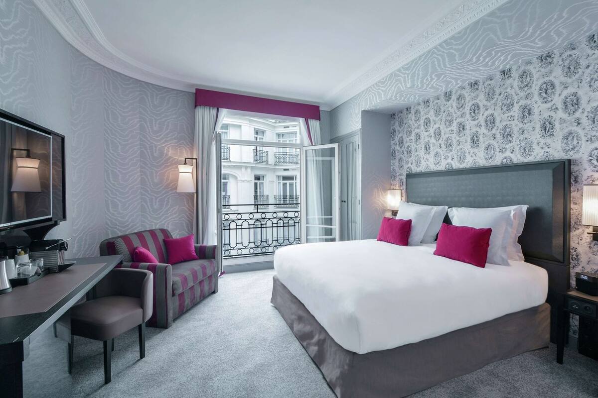 A hotel room with a bed, a desk, a chair, a TV, a balcony with a view of buildings, and patterned wallpaper.