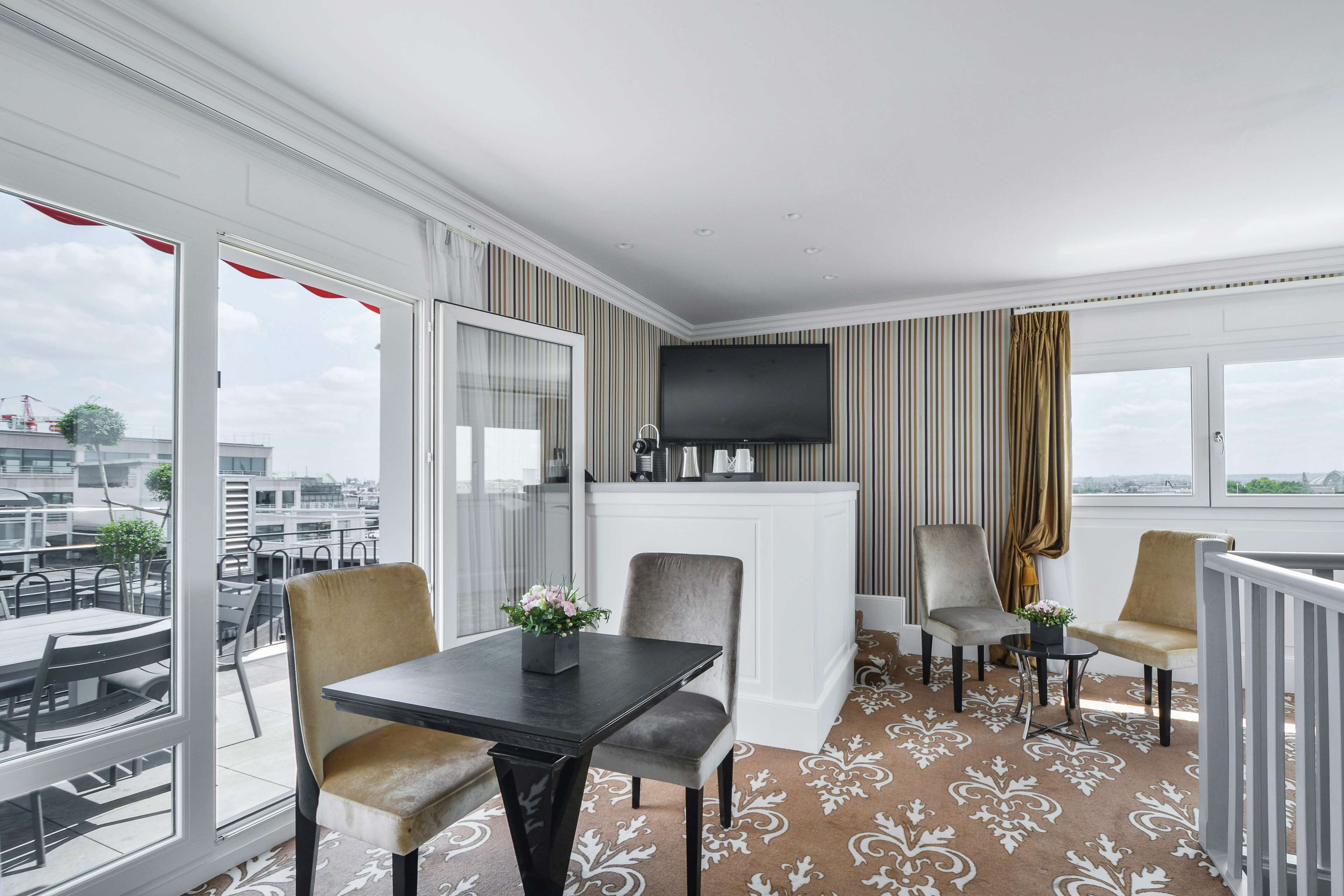 A hotel room with a balcony, a flat-screen TV, a dining table with chairs, and a patterned carpet.