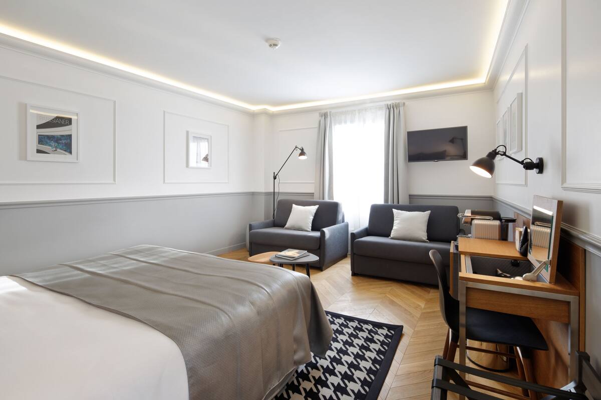 Moderni hotellihuone, jossa on suuri sänky, kaksi nojatuolia, työpöytä ja litteänäyttöinen televisio.