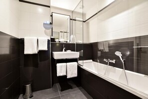 A modern bathroom with a black and white tiled design, a white bathtub, a glass shower enclosure, and a mirror above the sink. - XO Hotel (Paris)