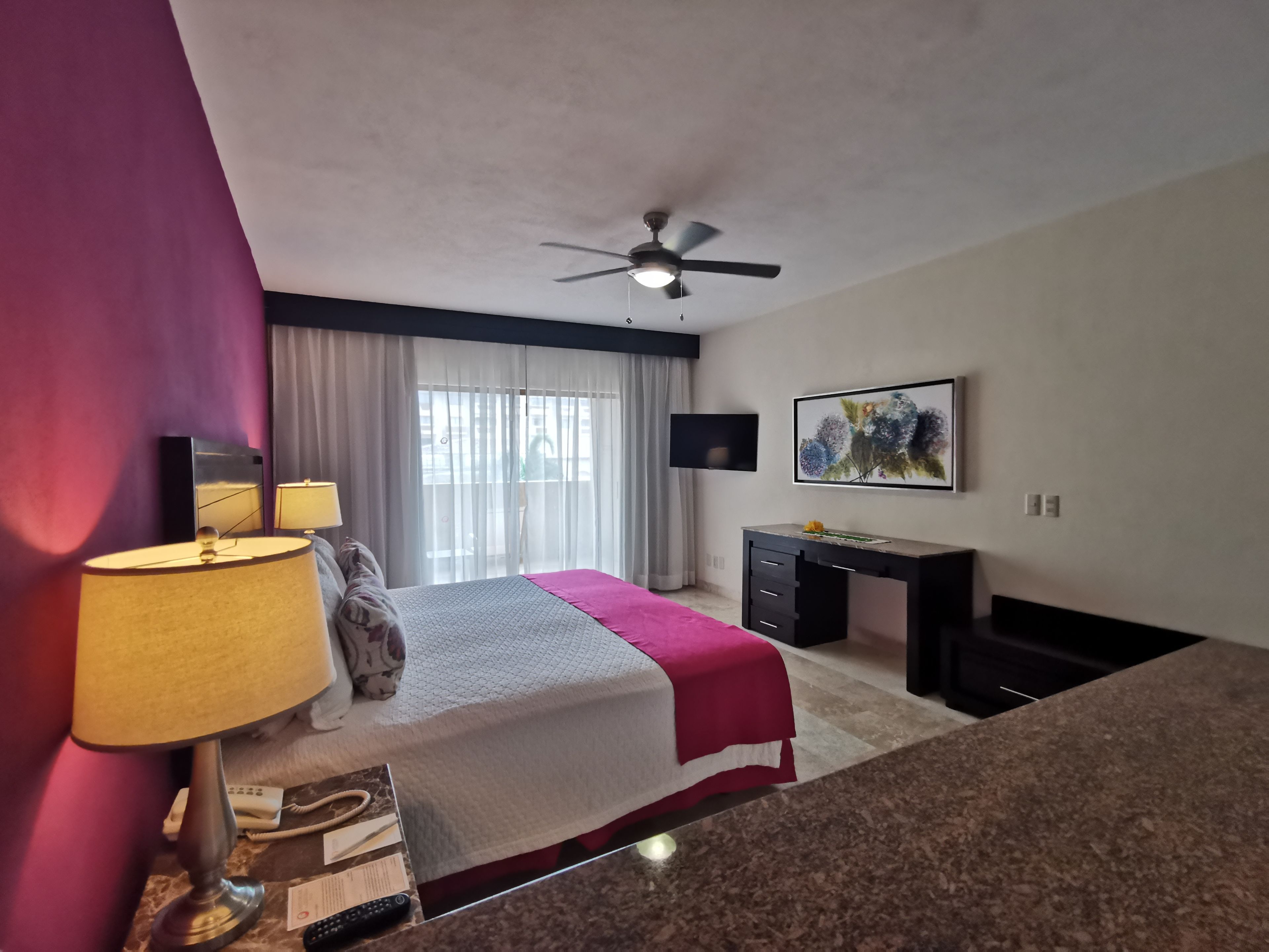 A hotel room with a bed, a desk, a ceiling fan, a TV, and a window with curtains.