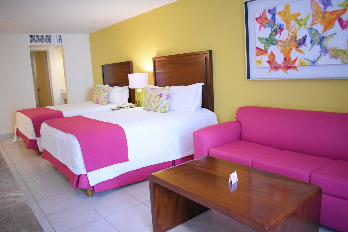 A hotel room with two beds, a pink sofa, and a wooden coffee table.