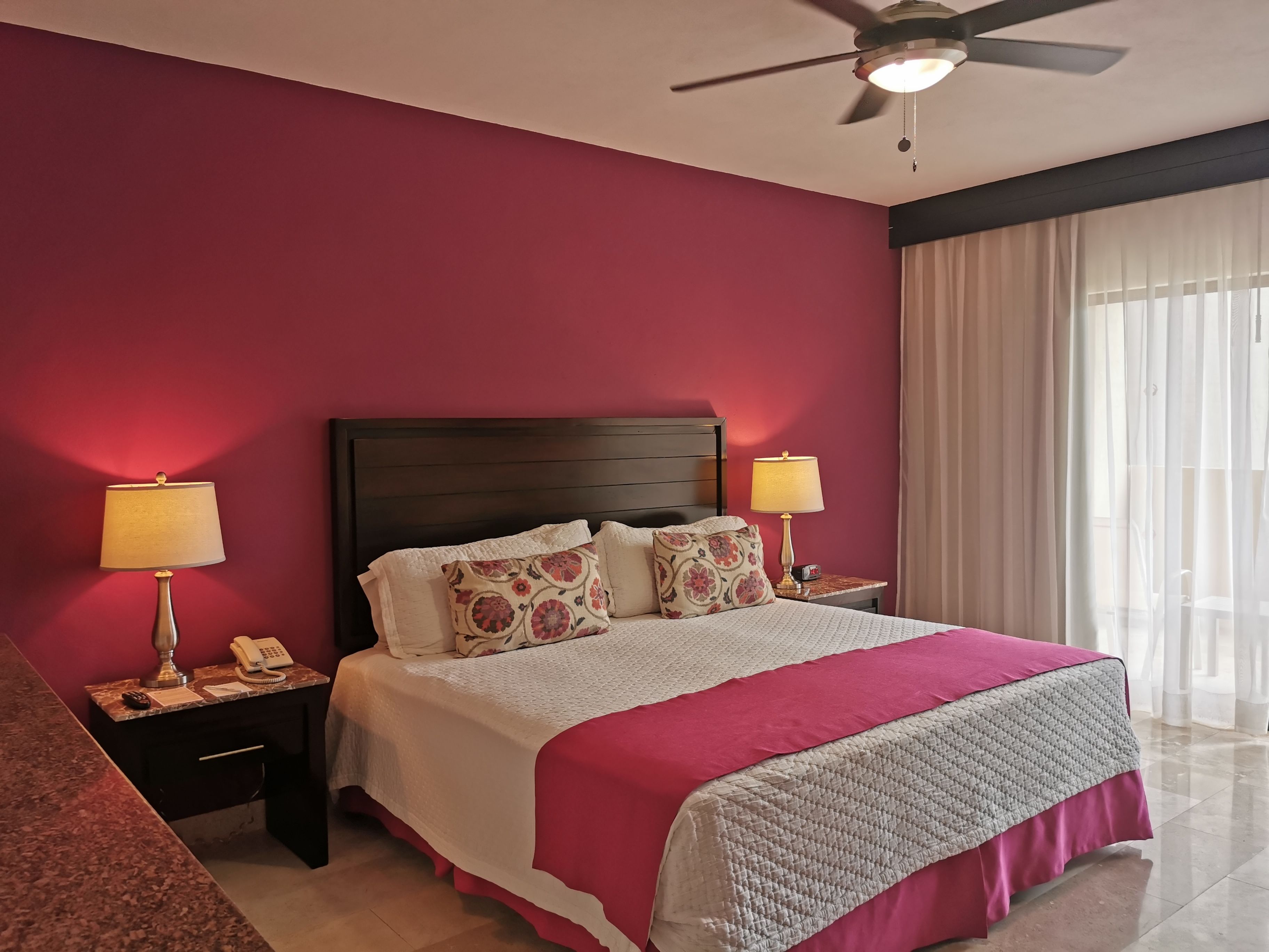 A hotel room with a large bed, two bedside tables, a ceiling fan, and a window with sheer curtains.