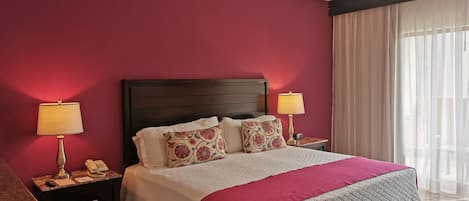 A hotel room with a large bed, two bedside tables, a ceiling fan, and a window with sheer curtains.