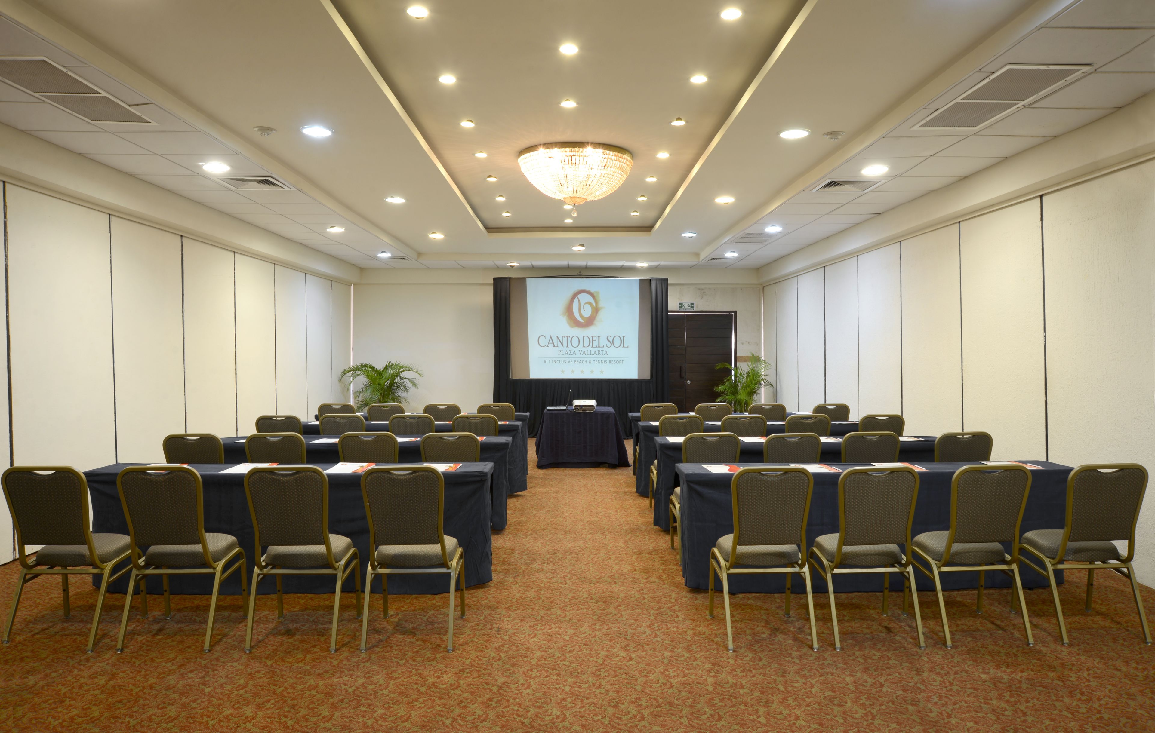 A conference room with a projector screen displaying 'CANTO DEL SOL PLAZA VALLARTA' at the front. Rows of tables with chairs are arranged for meetings.