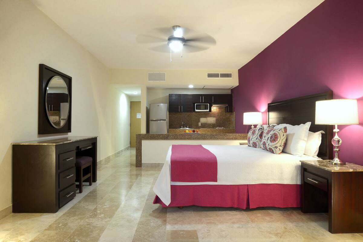 A hotel room with a bed, bedside table, nightstand, and a kitchenette area.
