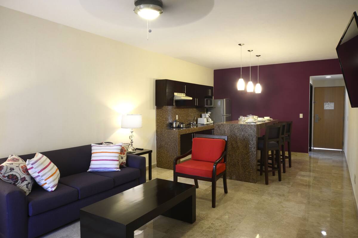 A modern living room with a dark sofa, a red chair, a coffee table, and a kitchen area with a microwave, stove, and bar counter.