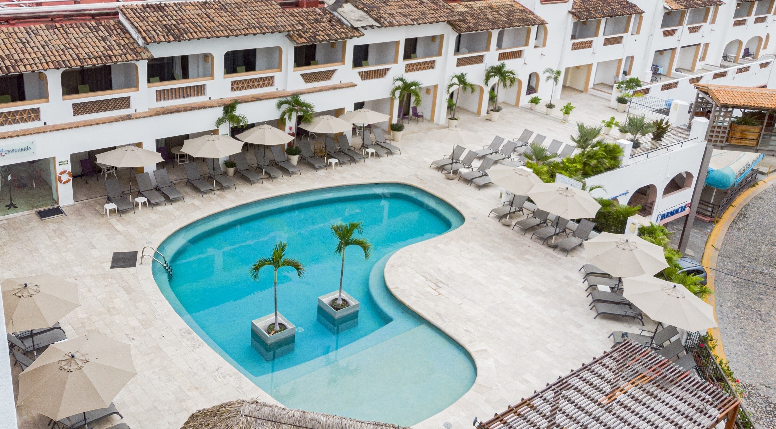 Un espace piscine agrémenté de chaises longues et de parasols, entouré d’un bâtiment doté de nombreux balcons et d’un toit en tuiles.