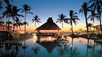 A resort pool with palm trees, a thatched-roof structure, and lounge chairs.