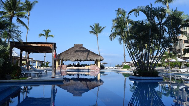 A resort pool with a thatched hut, palm trees, and lounge chairs. - Canto del Sol Puerto Vallarta All Inclusive (Puerto Vallarta)