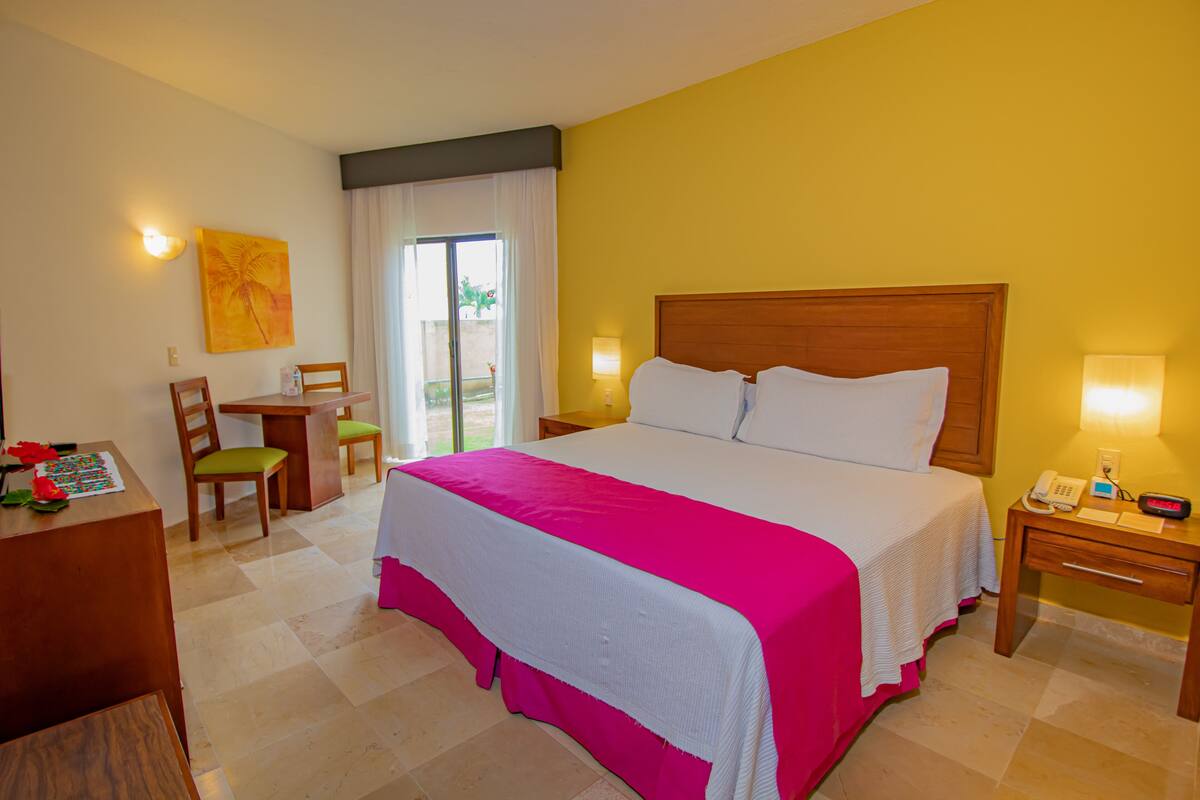 A hotel room with a large bed, a desk with a chair, a small table, and a nightstand with a lamp and phone.