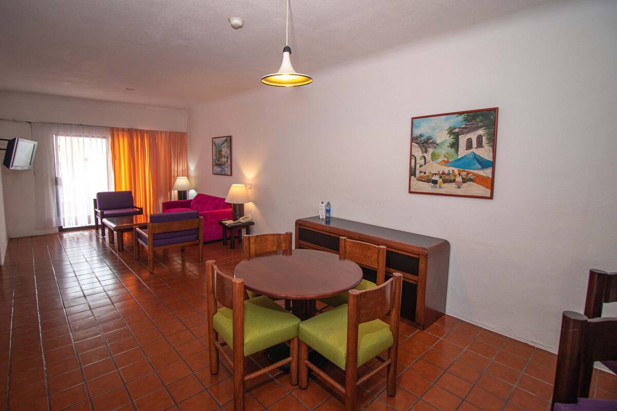 A dining area with a round table and four chairs, a bench, a TV, and a painting on the wall.