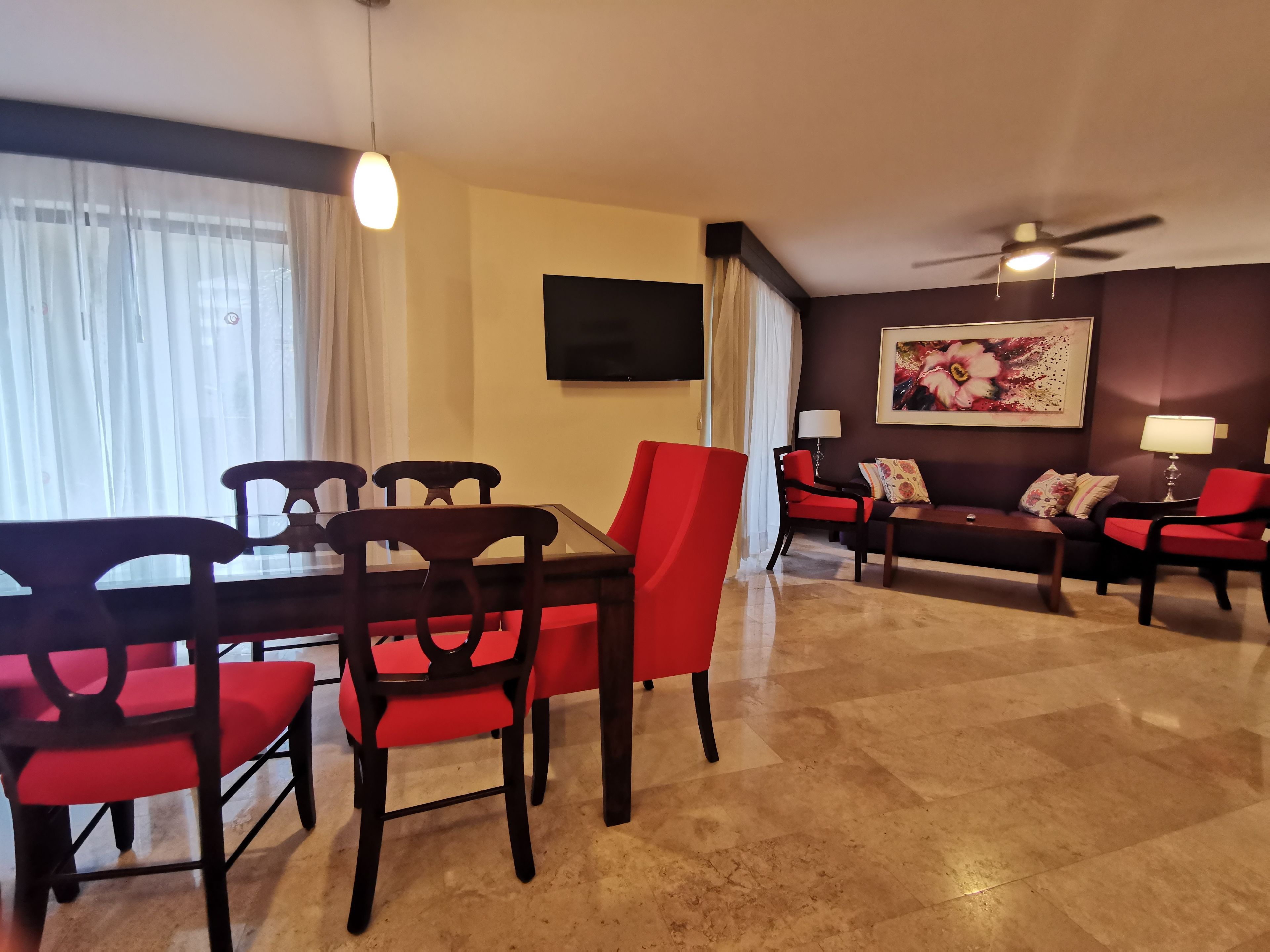 Un coin repas avec une table et des chaises, un ensemble de canapés, un téléviseur fixé au mur et un ventilateur de plafond.