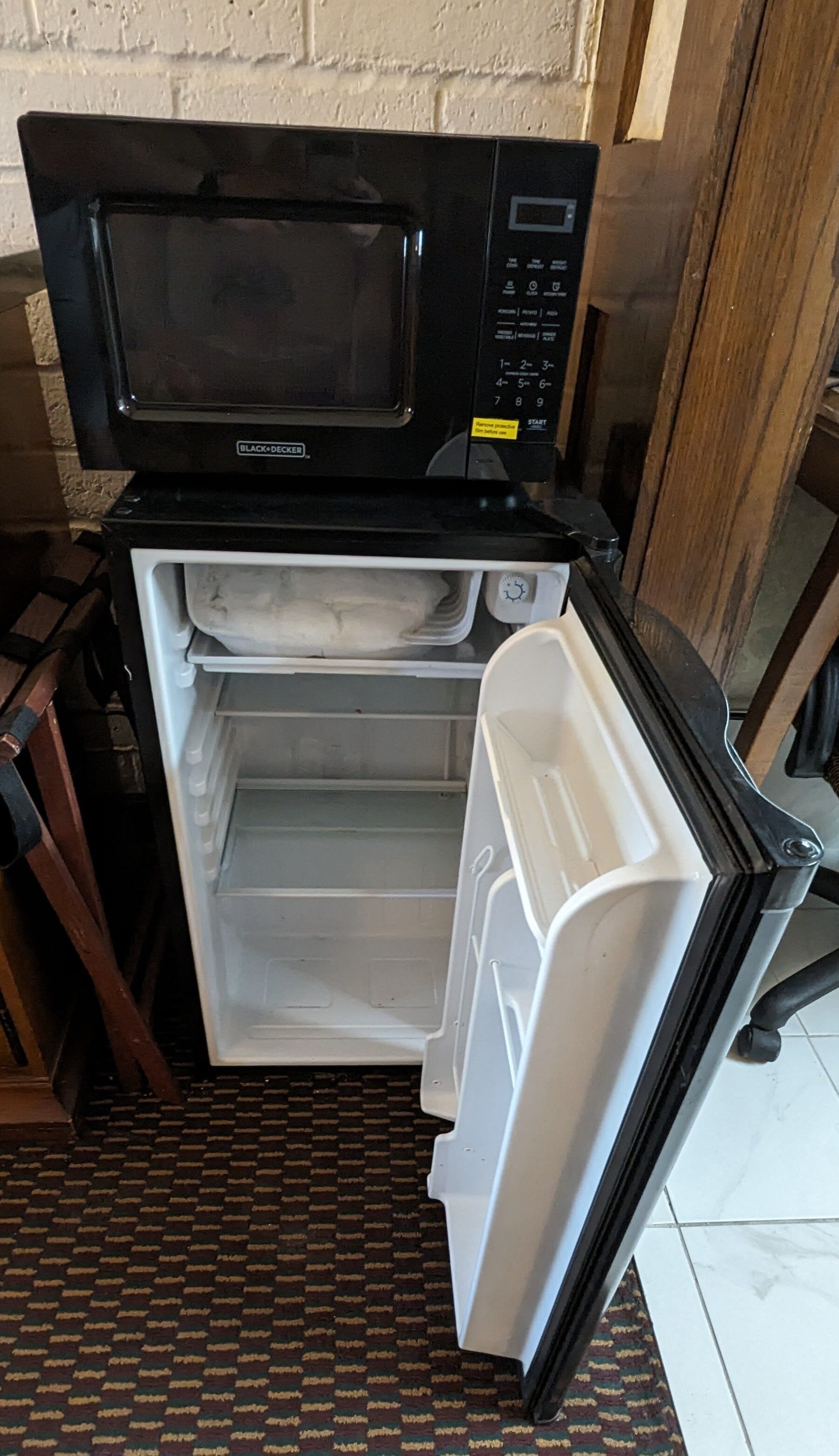 A compact black microwave with a digital display and control panel, a small refrigerator with a freezer compartment, and a patterned floor.