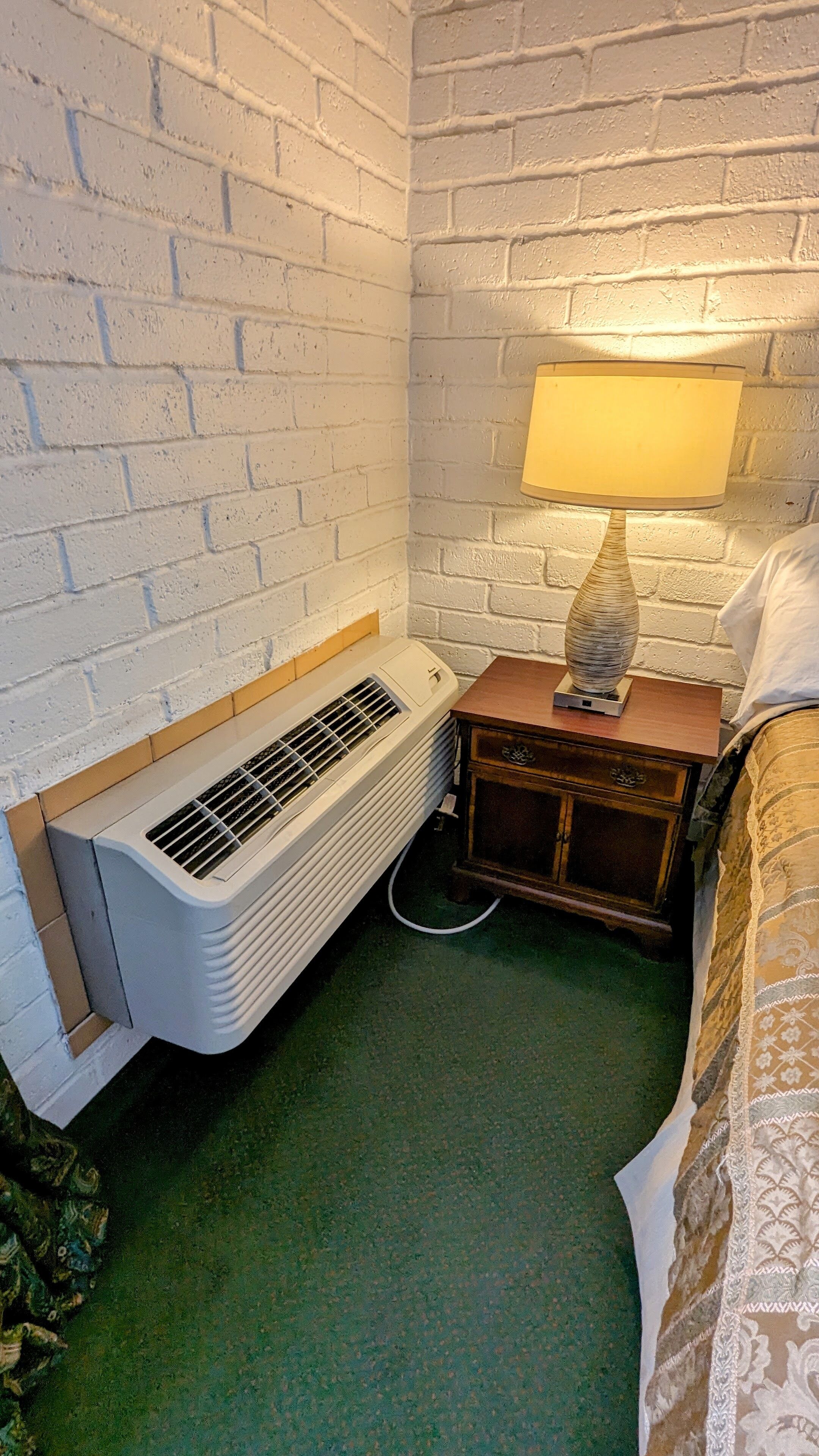 A corner room with a white brick wall, a heater, a wooden nightstand with a lamp, and a bed with a patterned bedspread.