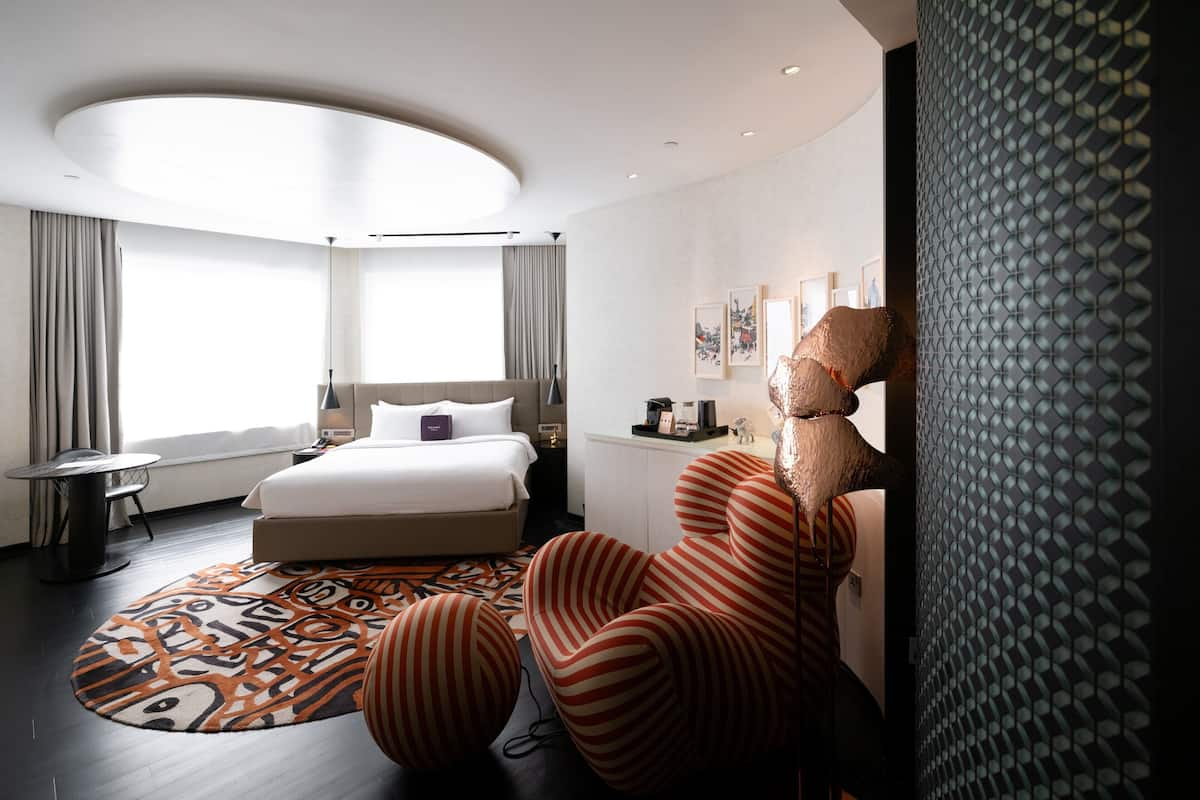 A modern hotel room with a large bed, a desk, a chair, and a patterned rug.