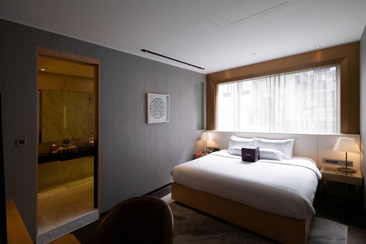 A hotel room with a bed, bedside tables, a window with curtains, a bathroom with a mirror, and a framed artwork on the wall.