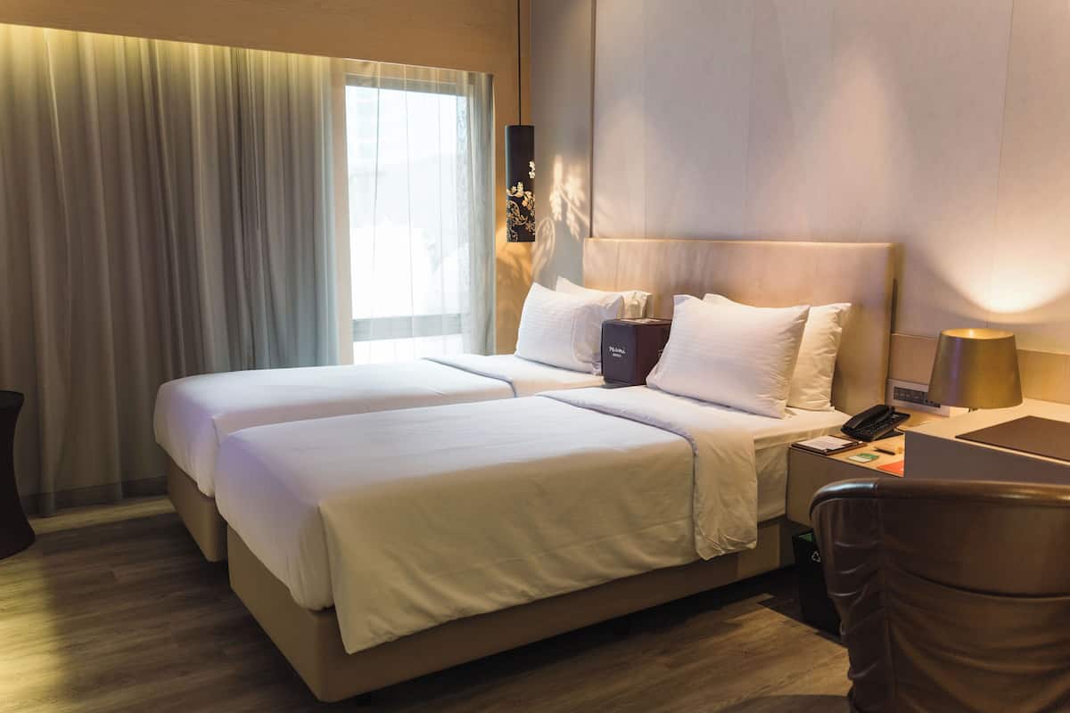A hotel room with two beds, a desk with a lamp, a phone, and a trash bin.