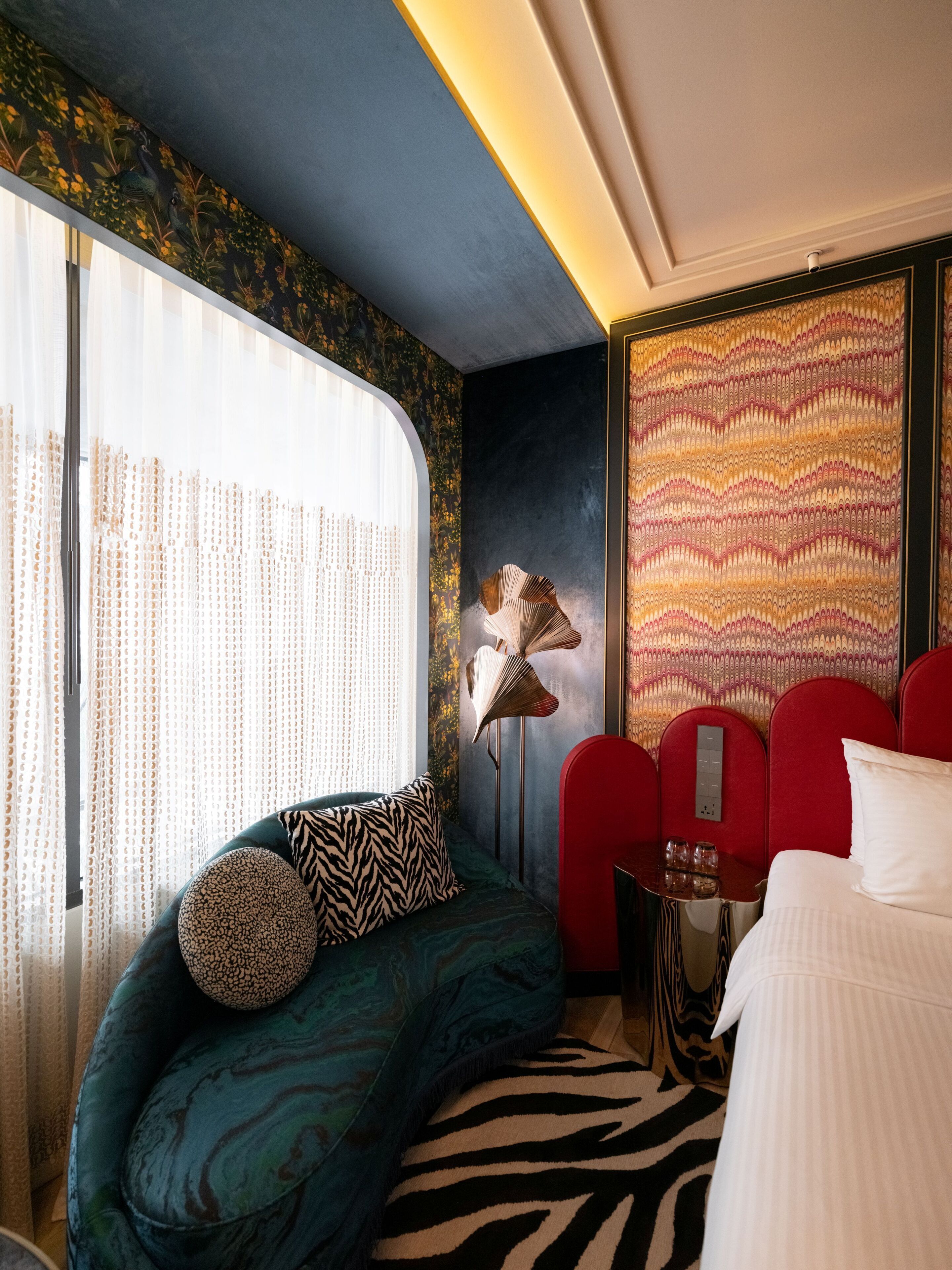 A hotel room with a sofa, a bed, a zebra-patterned rug, a red chair, and a wall with a decorative pattern.
