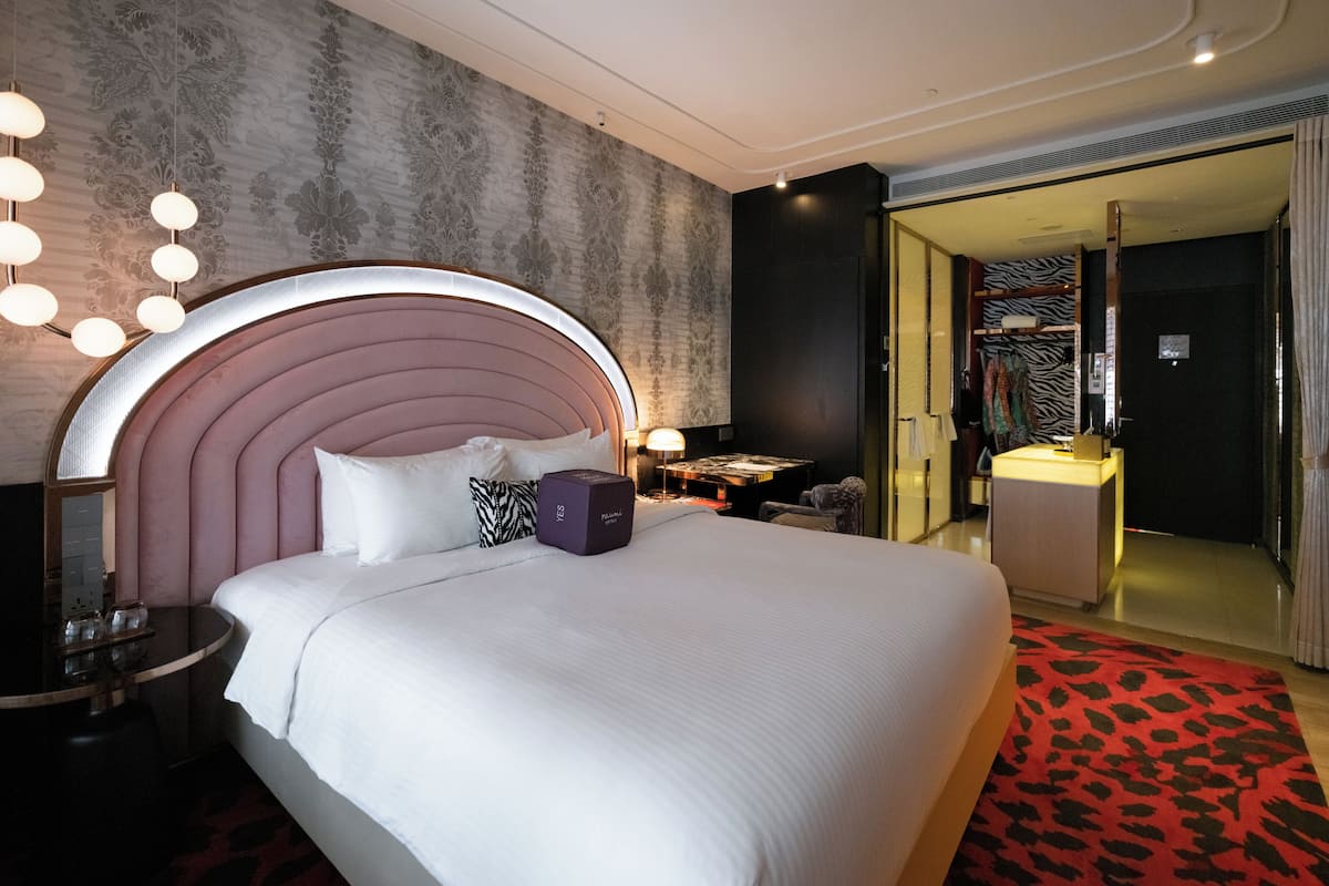 A hotel room with a large bed, a bedside table, a lamp, a wardrobe, and a bathroom visible through an open door.